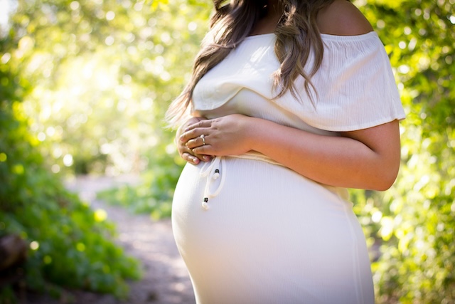 Pregnant woman standing in a garden wearing a white dress holding onto her belly